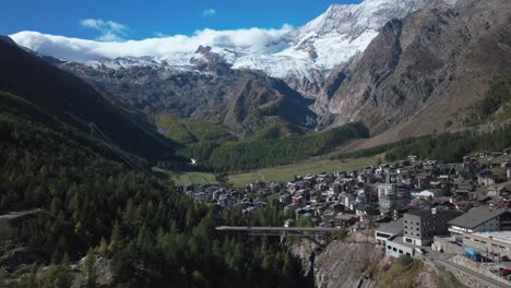 Sunny-stunning-Saastal-Saas-Fee-village-town-Switzerland-aerial-drone-beautiful-Fall-Autumn-Swiss-Alps-mountain-peaks-glacier-surrounding-city-buildings-bridge-Zermatt-The-Matterhorn-backwards-motion