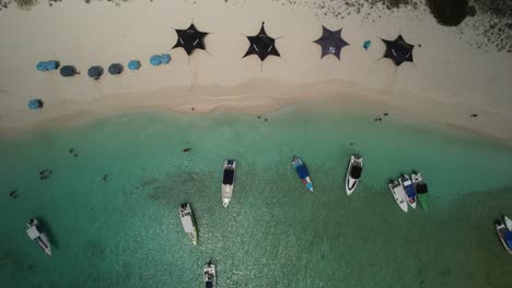 Boats-docked-at-a-pristine-sandy-beach-with-turquoise-waters-and-beach-umbrellas,-aerial-view