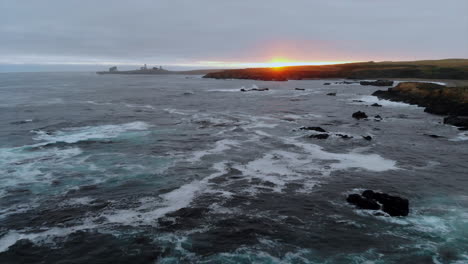 Roiling-sea-and-crashing-waves,-drone-rises-from-sea's-surface-to-pull-back-at-sunset,-Piedras-Blancas-Lighthouse-in-distance-on-Central-California-coast,-good-for-closing-credits,-4k-Pro-Res-422-HQ