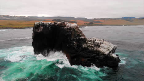 Crashing-Pacific-Ocean-waves-on-bird-rookery-rock-off-Central-California-Coast,-drone-flies-over-toward-beautiful-San-Simeon-cliffs-with-Hearst-Castle-in-distance-along-Highway-1,-4k-Pro-Res-422HQ