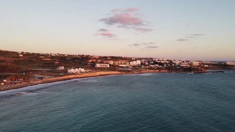 Aerial-view-of-Kissonerga-village-in-Cyprus-at-sunset,-with-golden-hues-illuminating-the-buildings-and-beach