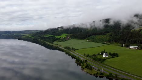 Luftaufnahmen-Zeigen-Die-Vingrom-Kirche,-Den-Mjøsa-See-Und-Die-Nahe-Gelegene-Autobahn-Vor-Der-Kulisse-üppiger-Grüner-Felder-Und-Nebelbedeckter-Hügel-In-Norwegen