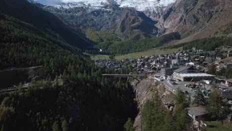 Sunny-stunning-Saastal-Saas-Fee-village-town-Switzerland-aerial-drone-beautiful-Fall-Autumn-Swiss-Alps-mountain-peaks-glacier-surrounding-city-buildings-bridge-Zermatt-The-Matterhorn-forwards-pan-up
