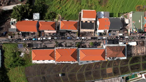 Top-down-view-of-heavy-traffic-with-cars-and-scooters-on-a-road-lined-with-buildings-with-orange-roofs-in-Canggu,-surrounded-by-green-rice-fields-and-other-structures