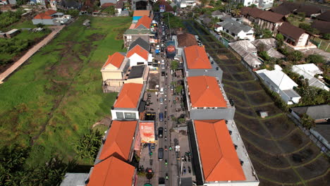 Aerial-view-of-heavy-traffic-along-a-narrow-street-in-Canggu,-lined-with-buildings-with-red-roofs-and-surrounded-by-green-fields-and-residential-areas