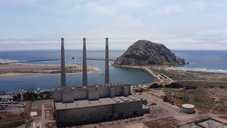 Wide-push-in-aerial-shot-of-the-abandoned-Morro-Bay-Power-Plant-with-its-famous-three-smokestacks-in-front-of-Morro-Rock-in-Morro-Bay,-California