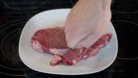 Hand-rubbing-salt-and-pepper-into-raw-entrecote-on-white-plate,-preparing-for-cooking