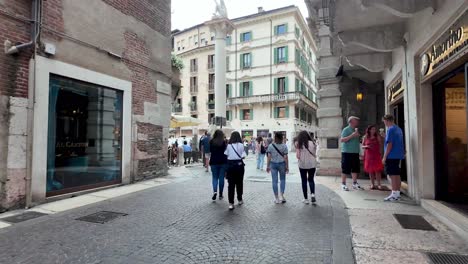 Caminando-Por-La-Calle-Llena-De-Gente-Hacia-La-Apertura-Y-La-Plaza,-Una-Famosa-Piazza-Delle-Erbe-Con-Estatuas,-Torres,-Mercado,-Cafés,-Tiendas,-Etc.