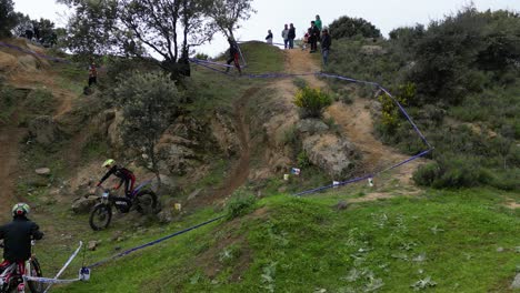 Drohnenflug-Bei-Einem-Motocross-Test-Im-Freien,-Bei-Dem-Der-Pilot-Auf-Der-Mit-Blauem-Band-Markierten-Strecke-In-Aktion-Ist,-Auf-Und-Ab-Geht,-Gut-Ausgeführt,-Es-Gibt-Leute,-Die-Zuschauen-Und-Spaß-Haben