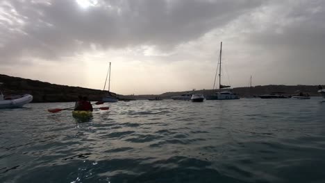 Kayaker-couple-during-sunset-at-Blue-Lagoon,-Comino-with-moored-boats-and-Gozo-in-the-background-at-Malta