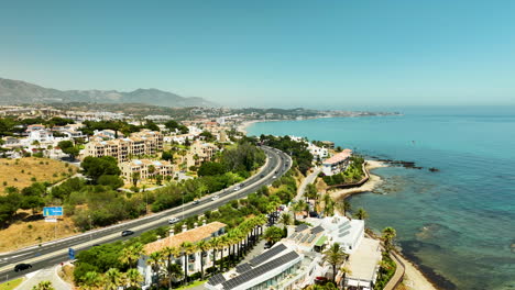 Aerial-shot-of-coastal-road,-seaside-buildings,-and-turquoise-sea-with-clear-blue-sky-in-Mijas,-Spain