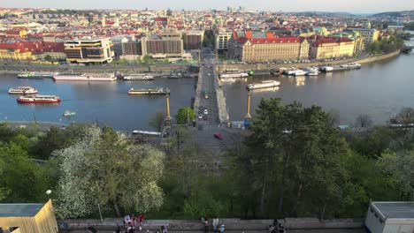 Prague-metronome-Aerial-drone-descends-letna-park-as-tourists-look-out-over-city-skyline-over-bridge-at-sunset,-czechia,-czech-republic