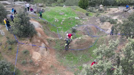 Filmación-Con-Drone-Donde-Vemos-Una-Prueba-De-Motocross-Donde-El-Asesor-Del-Piloto-Tiene-Que-Darle-Una-Mano-Para-Superar-El-Obstáculo-Dentro-Del-Circuito-Marcado,-Hay-Espectadores-Disfrutando-Del-Evento