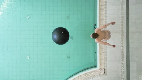 Woman-jumps-onto-and-grabs-hold-of-floating-exercise-ball-in-pool,-overhead-view