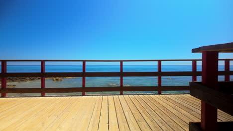 Sonniges-Deck-Mit-Blick-Auf-Den-Klaren-Blauen-Ozean-Mit-Einem-Holzgeländer-Und-Ruhigem-Wasser-Im-Hintergrund