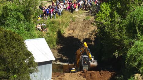 Aerial-shot-showcasing-an-excavator-digging-dirt-while-a-crowd-of-people-watches-in-Chiloe,-Chile