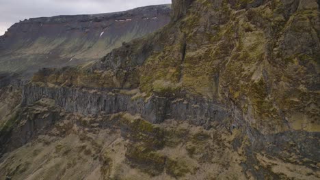 Aerial-cinematic-shot-of-mountain-rock-formations-and-steep-cliffs,-with-melting-snow,-on-a-moody-evening-in-Iceland