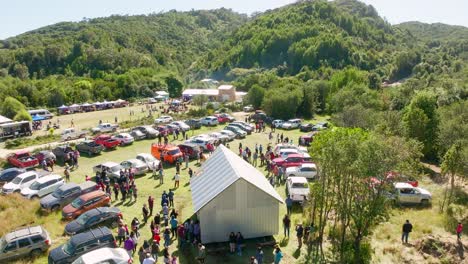 Aerial-scenic-outdoor-event-with-crowds-gathering-around-tents-and-parked-cars-in-a-lush,-green-natural-area-surrounded-by-trees-and-hills-under-a-clear-sky