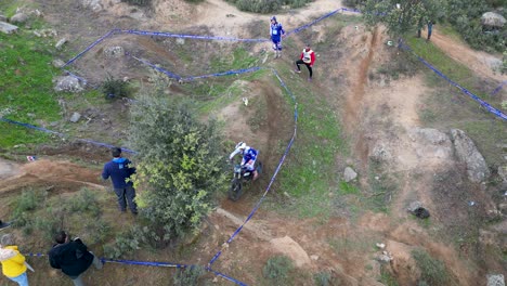 Drone-flight-in-an-outdoor-motocross-competition-where-the-pilot-is-in-action-on-the-circuit-marked-with-blue-tape,-his-advisor,-the-organization's-judges-and-spectators