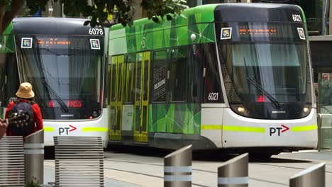 Trams-glide-along-the-tramway-through-the-bustling-downtown-Melbourne-Bourke-Street-Mall,-with-pedestrians-crossing,-commuters-waiting-at-the-tram-stop,-showcasing-the-city's-dynamic-urban-lifestyle