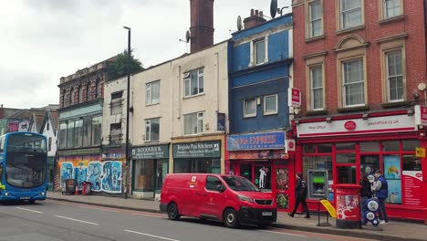 High-street-view-of-shops,-houses-and-traffic-with-Royal-Mail-delivery-van,-Post-Office-and-double-decker-bus-in-Stokes-Croft,-Bristol-City,-England-UK