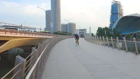 Young-Man-Riding-Bicycle-Across-Bridge-Near-City-Buildings-In-The-Morning