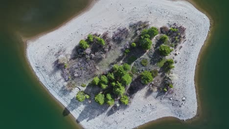 Drone-rises-above-sandy-island-with-evergreen-trees-casting-longshadow-across-water