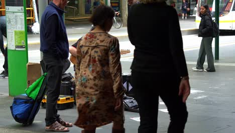 A-talented-musician-busks-on-Bourke-Street-Mall,-skilfully-playing-the-guitar-in-the-heart-of-Melbourne-city,-with-pedestrian-passing-by-in-the-scene