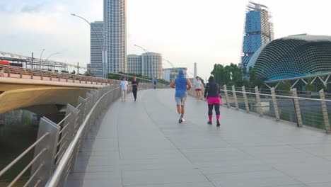 Hombres-Y-Mujeres-Atletas-Hacen-Jogging-Matutino-Moviéndose-A-Lo-Largo-De-Un-Camino-Concreto-En-Un-Moderno-Puente-Peatonal-En-La-Ciudad