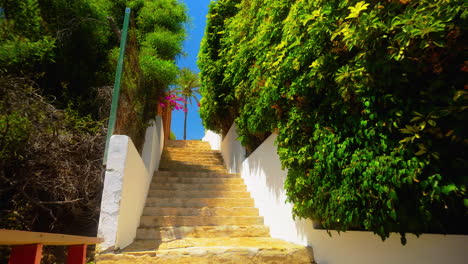 Sunny-stone-stairway-lined-with-lush-greenery-in-Mijas,-Spain,-leading-up-to-a-vibrant,-scenic-view