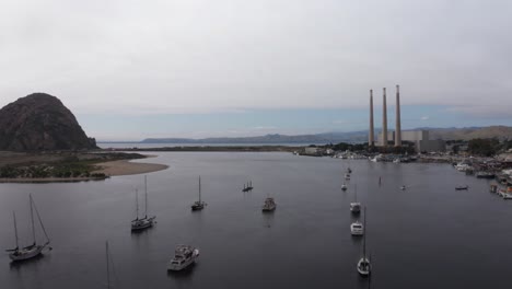 Toma-Aérea-Panorámica-Baja-De-Veleros-En-El-Puerto-De-Morro-Bay-Con-Morro-Rock-Y-Las-Tres-Pilas-De-La-Central-Eléctrica-Abandonada-De-Morro-Bay-En-La-Distancia