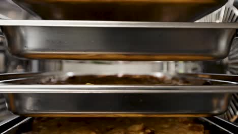 Industrial-kitchen-oven-filled-with-trays-of-food,-close-up-elevator-shot-of-bubbling-dishes