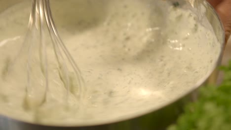 Close-up-of-whisk-blending-fresh-herbs-into-creamy-sauce-in-stainless-steel-bowl