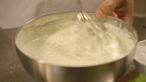 Close-up-of-a-chef-whisking-a-creamy-herb-sauce-in-a-stainless-steel-bowl,-panning-shot