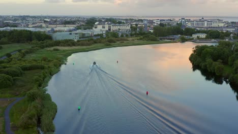 Speed-boat-drives-between-buoys-as-River-Corrib-water-reflects-golden-sunset-sky,-aerial