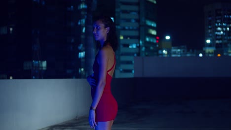 In-a-red-dress,-a-young-girl-stands-on-a-nighttime-rooftop,-with-city-buildings-as-her-backdrop