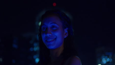 On-a-rooftop-under-the-stars,-a-girl-in-a-red-dress-stands-with-city-buildings-behind-her