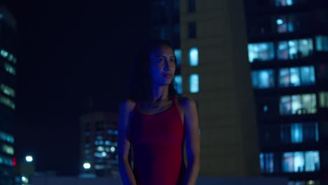 Dressed-in-red,-a-young-girl-stands-on-a-rooftop-at-night,-city-buildings-behind-her