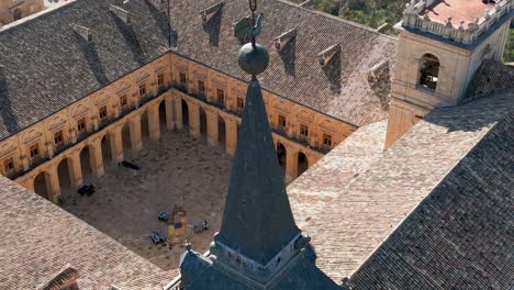 Vuelo-Ascendente-Visualizando-El-Hermoso-Claustro-Con-Sus-Arcos-Del-Monasterio-De-Ucles-Siglos-XVI-XVIII-Y-Su-Fuente-En-Medio-Del-Patio-Vemos-El-Techo-Negro-De-La-Torre