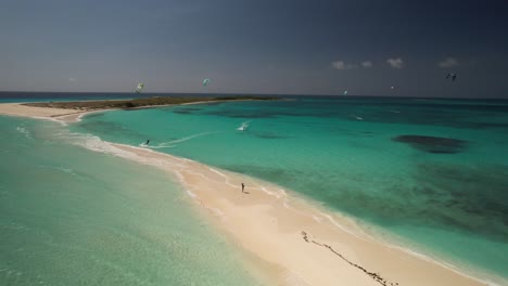 Kiteboarders-gliding-over-turquoise-waters-near-a-sandy-beach,-aerial-view