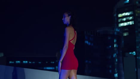 Wearing-a-red-dress,-a-young-girl-is-perched-on-a-rooftop-at-night,-city-buildings-forming-the-background