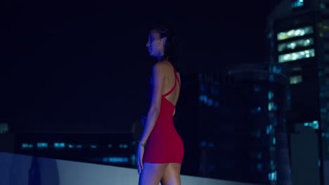 Con-Un-Vestido-Rojo,-Una-Joven-Está-Encaramada-En-Una-Azotea-Por-La-Noche,-Con-Los-Edificios-De-La-Ciudad-Formando-El-Fondo