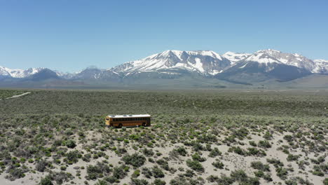 Yellow-school-bus-travels-across-a-vast-desert-with-snow-capped-mountains-in-the-distance