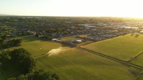 Un-Dron-Sobrevuela-Los-Verdes-Campos-De-Nueva-Zelanda,-Capturando-A-Un-Irrigador-En-Acción,-Conduciéndolo-Hacia-Una-Ciudad-Distante