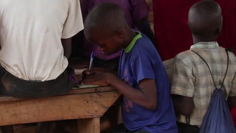 Kleiner-Junge-In-Einem-Blauen-Hemd-Konzentriert-Sich-Auf-Das-Schreiben-An-Einem-Holzschreibtisch,-Umgeben-Von-Anderen-Kindern-In-Uganda