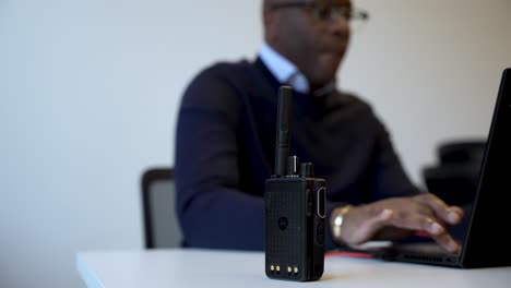 Close-up-of-a-walkie-talkie-on-a-table-with-a-blurred-businessman-working-on-a-laptop-in-the-background-in-an-office-environment