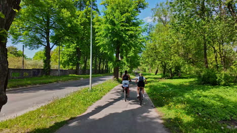 Two-white-women-driving-the-bicycle-in-a-green-park-in-spring-slowmotion-dolly-shot-following-persons
