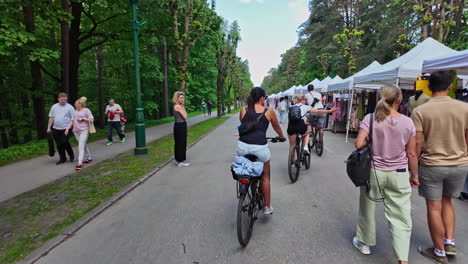 White-people-with-children-on-a-market-street,-walking-and-two-white-women-and-a-man-riding-bicycle-passing-the-tents-slowmotion-following-them