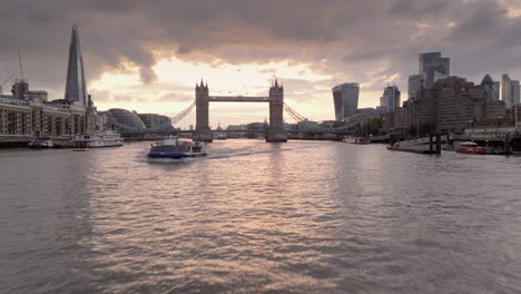 Low-drone-dolly-past-ferry-on-River-Thames-toward-London-Tower-Bridge-at-sunset