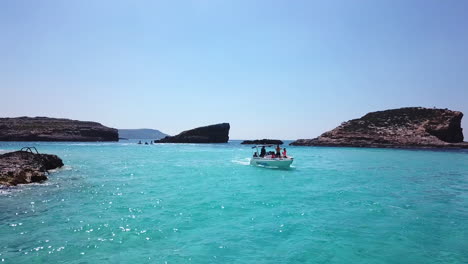 Eine-Drohne-Fliegt-Rückwärts-Und-Nimmt-Ein-Kleines-Boot-Auf,-Das-Entlang-Der-Blauen-Lagune-Auf-Der-Insel-Comino-In-Malta-Fährt